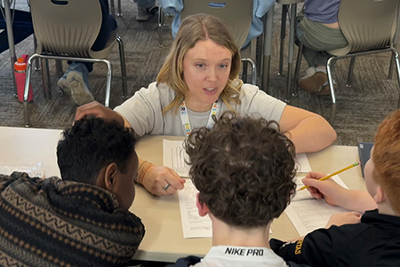 INSPIRE staff squatting in front of three students at table looking at worksheet