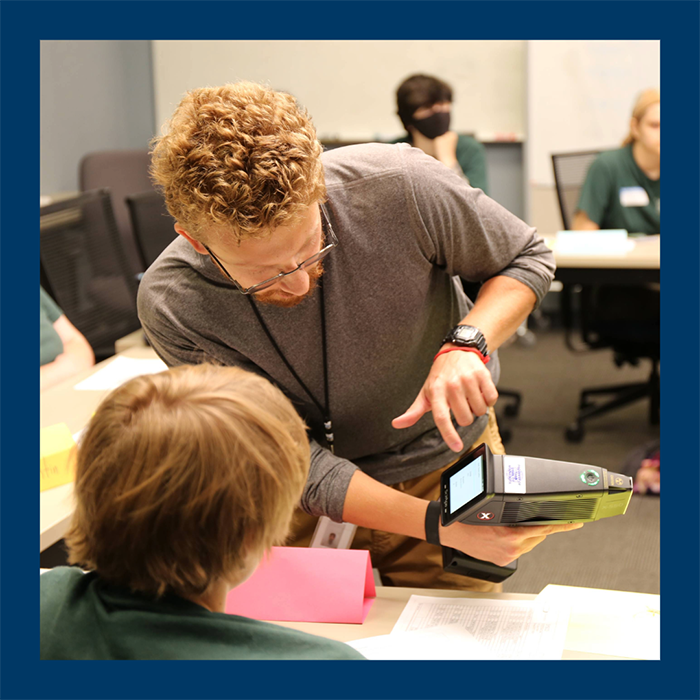 adult with ID badge stands next to sitting kid and points to screen on handheld machine